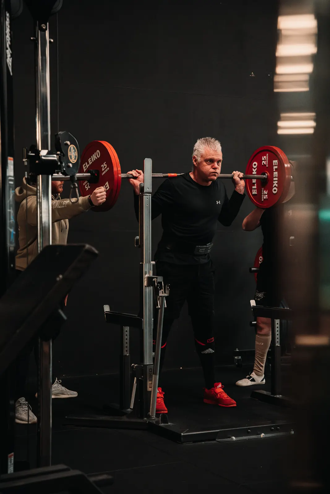 Lifter met grijs haar staat geconcentreerd onder de stang in het rack, rode Eleiko-schijven aan weerszijden