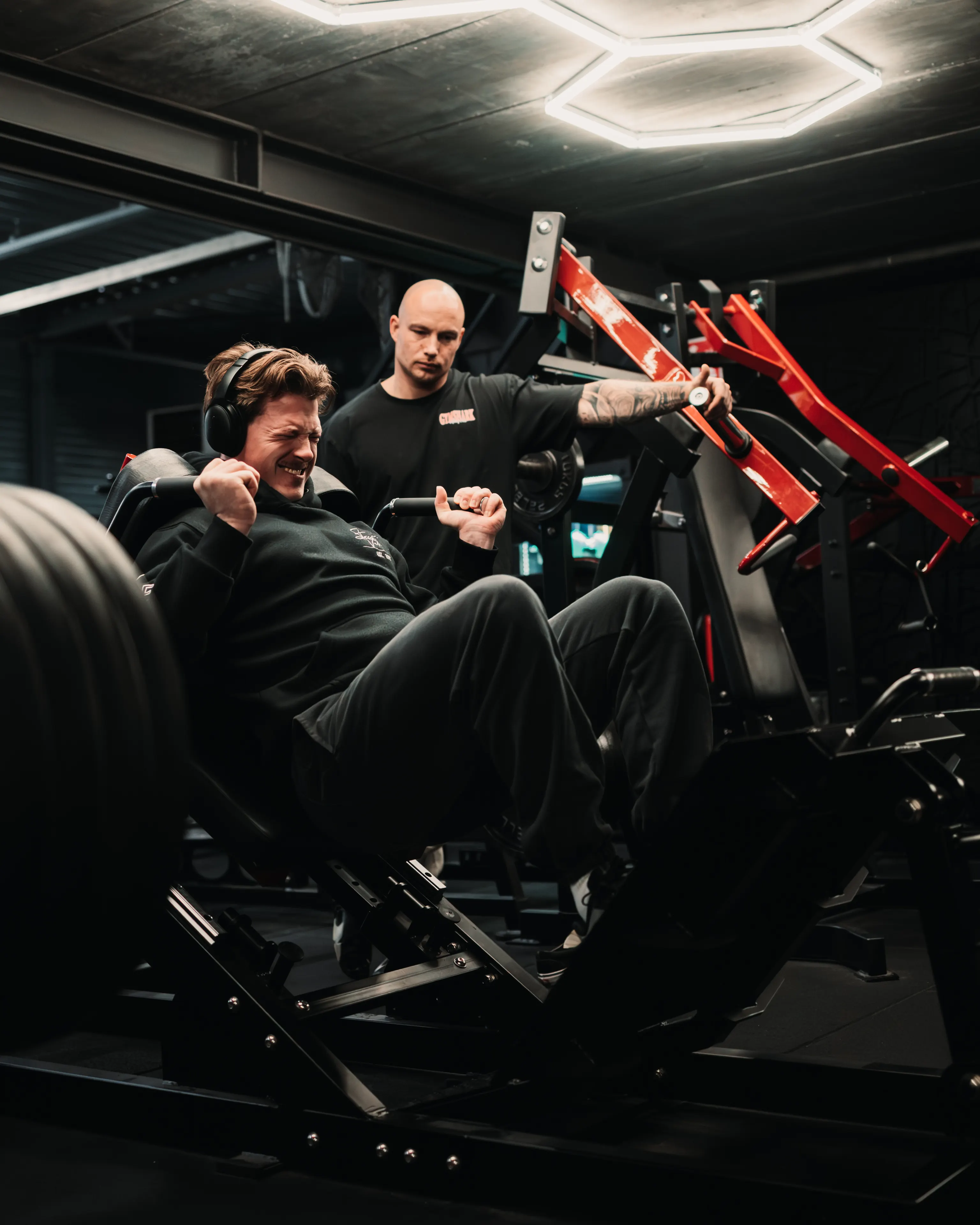 Lifter drukt een zwaar beladen pendulum squat weg terwijl een coach meekijkt